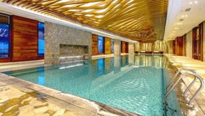 An indoor swimming pool with a stone wall and wooden paneling, surrounded by seating areas and a patterned ceiling. - Genting Grand Chongli (Zhangjiakou)