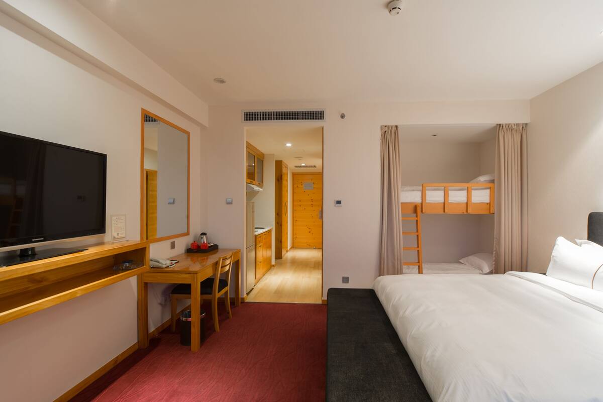 A hotel room with a TV, desk, bunk bed, and a small kitchenette.
