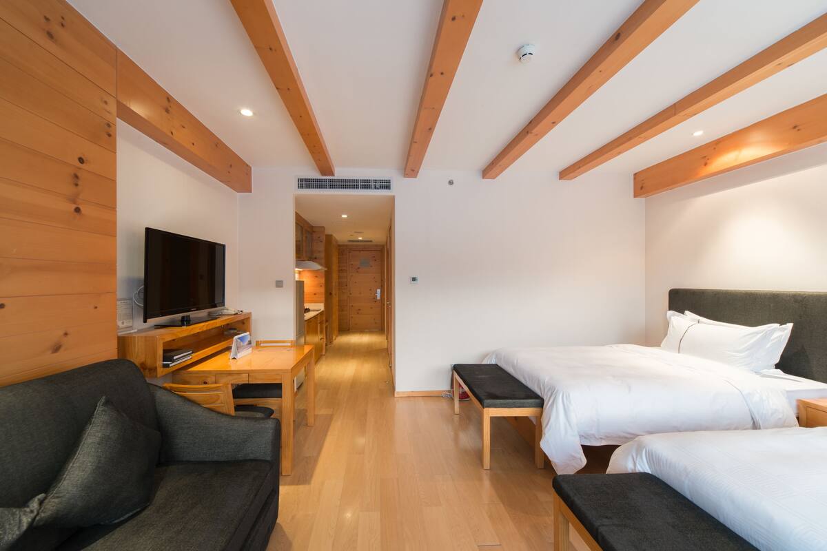 A modern hotel room with wooden beams, a flat-screen TV, a sofa, two beds, and a desk.