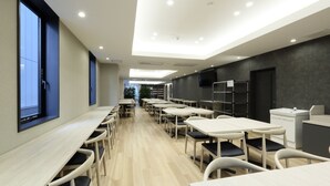 Een moderne eethoek met meerdere tafels en stoelen, een groot raam links en een keuken aan de rechterkant.
