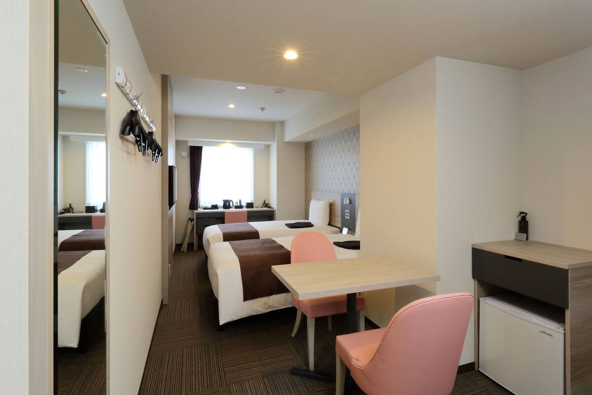 A hotel room with a bed, a desk, a chair, a small refrigerator, and a mirror on the wall.