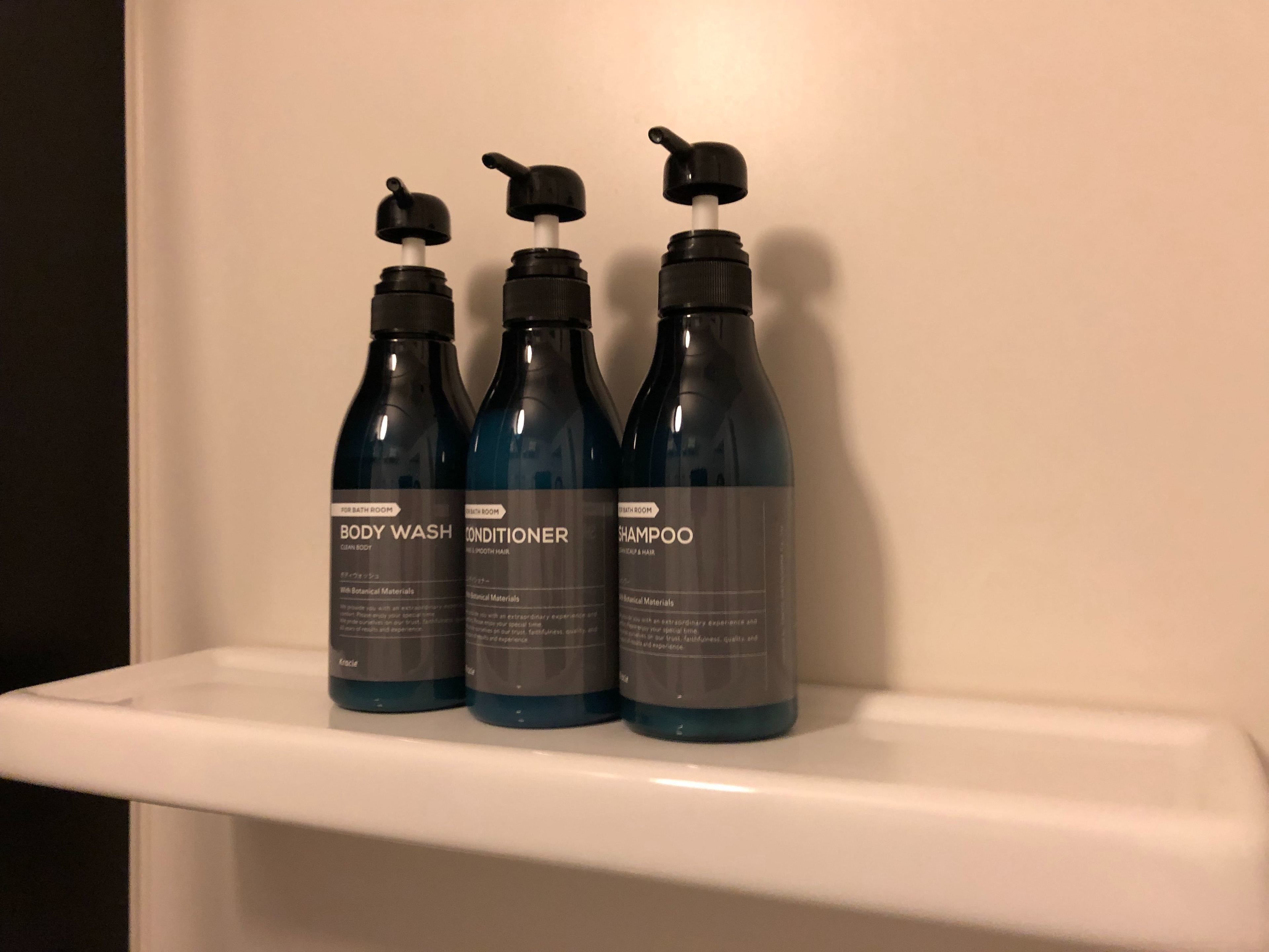 Trois bouteilles de gel douche, d’après-shampoing et de shampoing sont posées sur une étagère blanche.