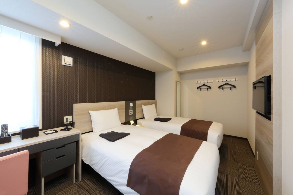 A hotel room with two beds, a desk, a chair, a TV, and a window.