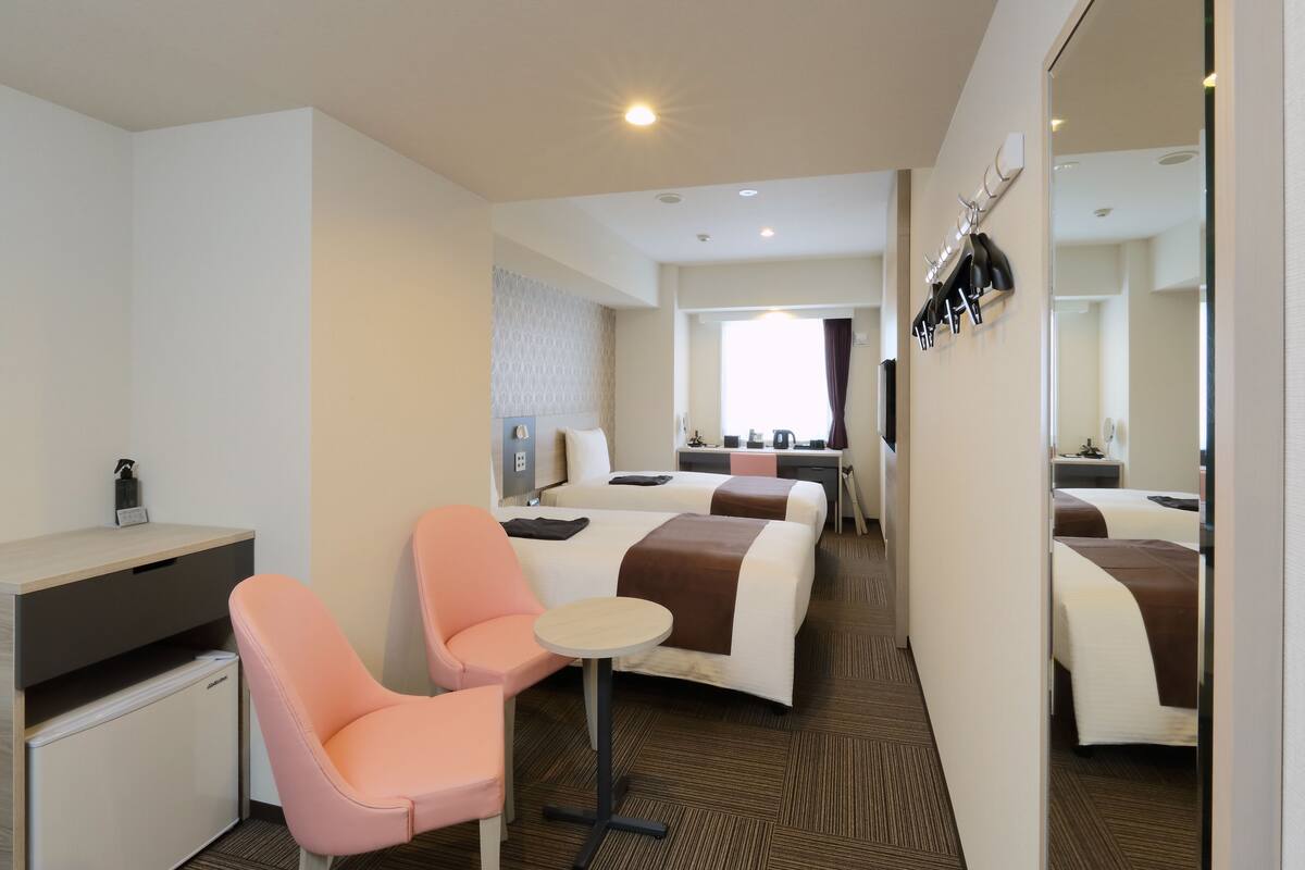 A hotel room with a desk, two beds, a small table, a chair, and a mini-fridge.