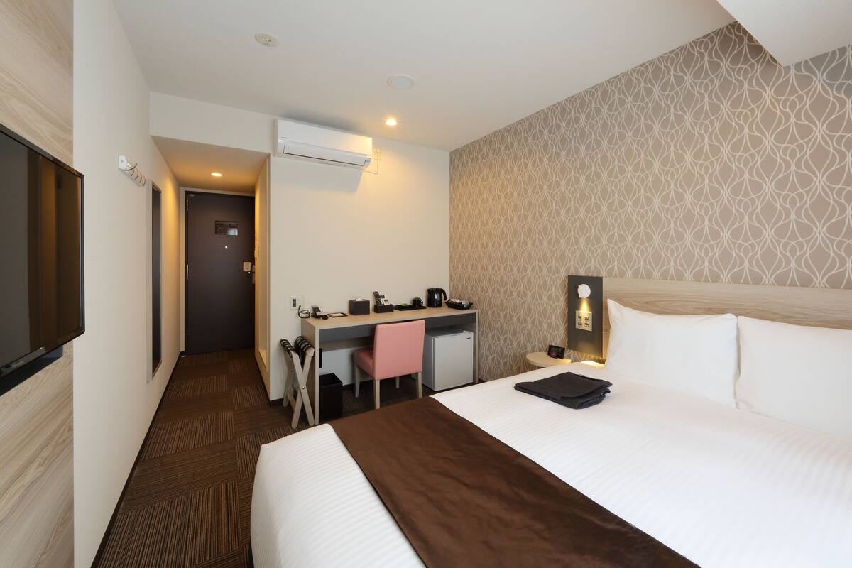 A hotel room with a large bed, a desk with a chair, a small desk, an air conditioning unit, and a television.