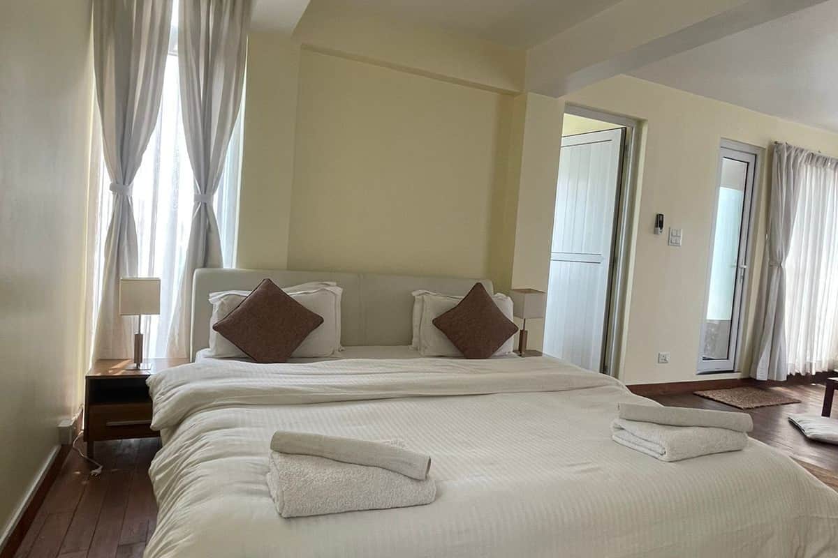 A large, neatly made bed with white linens and brown pillows in a hotel room with wooden flooring and a large window with curtains.