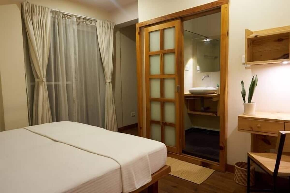 A bedroom with a bed, a wooden wardrobe, a desk, and a potted plant.