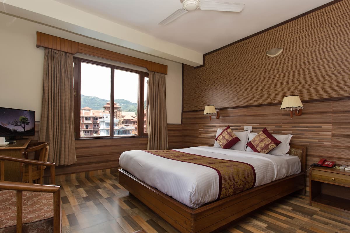 A hotel room with a large bed, wooden flooring, a desk with a chair, a TV, a window with curtains, and a wall-mounted lamp.