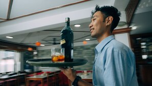 Une personne tenant un plateau sur lequel se trouvent une bouteille et un verre de jus d’orange, dans un restaurant.