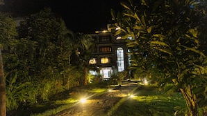 Une vue nocturne d’un bâtiment entouré d’une végétation luxuriante, avec un chemin bien éclairé menant à l’entrée.