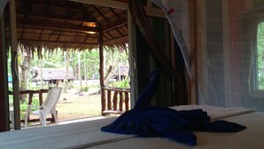 A room with a bed, a chair, and a view of a thatched-roof structure outside.