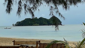 A beach with a bamboo structure in the foreground, a small boat on the water, and a forested island in the distance.