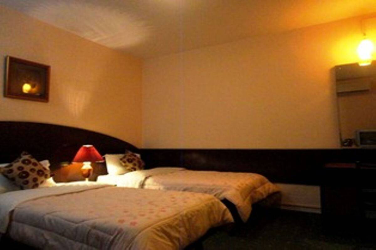 A hotel room with two beds, a nightstand with a lamp, a wall-mounted air conditioner, and a framed picture on the wall.