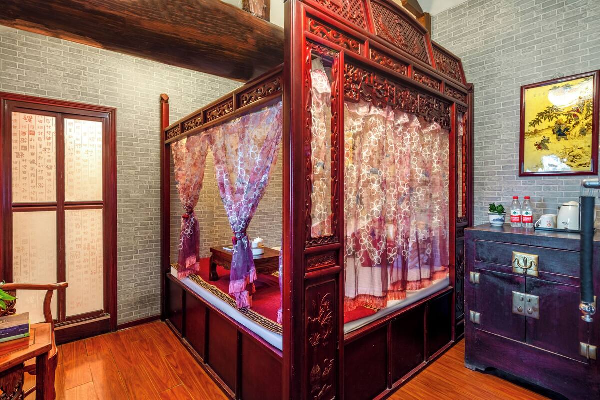 A traditional room with a wooden bed, a small table with books, and a cabinet with bottles.