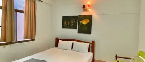 Un dormitorio con una cama de madera, dos almohadas, una mesita de noche, un ventilador verde y blanco, una lámpara de techo y dos marcos con cuadros colgados en la pared.