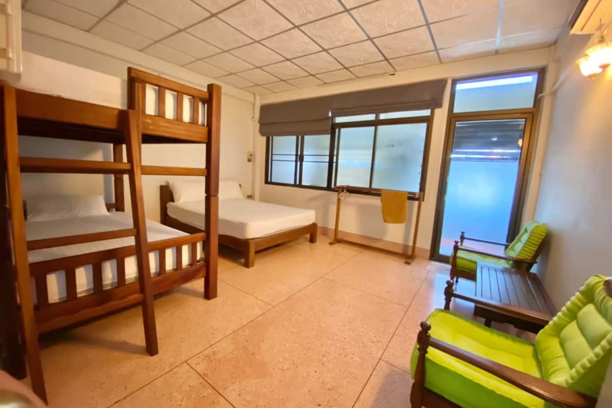 Une chambre avec des lits superposés, un lit simple, une fenêtre avec des stores, une chaise verte et une petite table.
