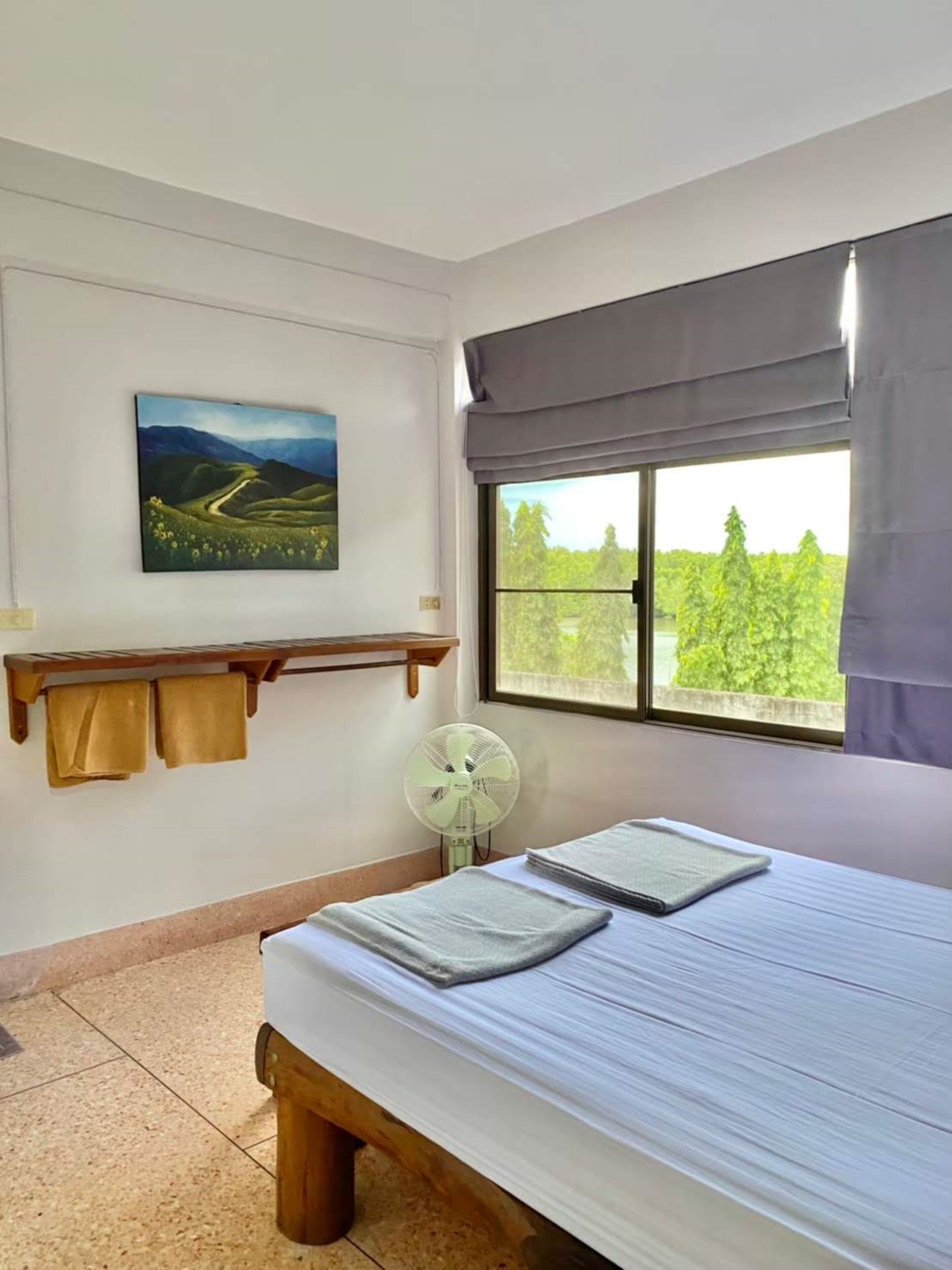 Um quarto com cama, janela com cortinas, ventilador fixado na parede, prateleira com toalhas e uma pintura.