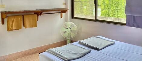 Un dormitorio con cama, ventana con cortinas, ventilador de pared, estante con toallas y un cuadro.