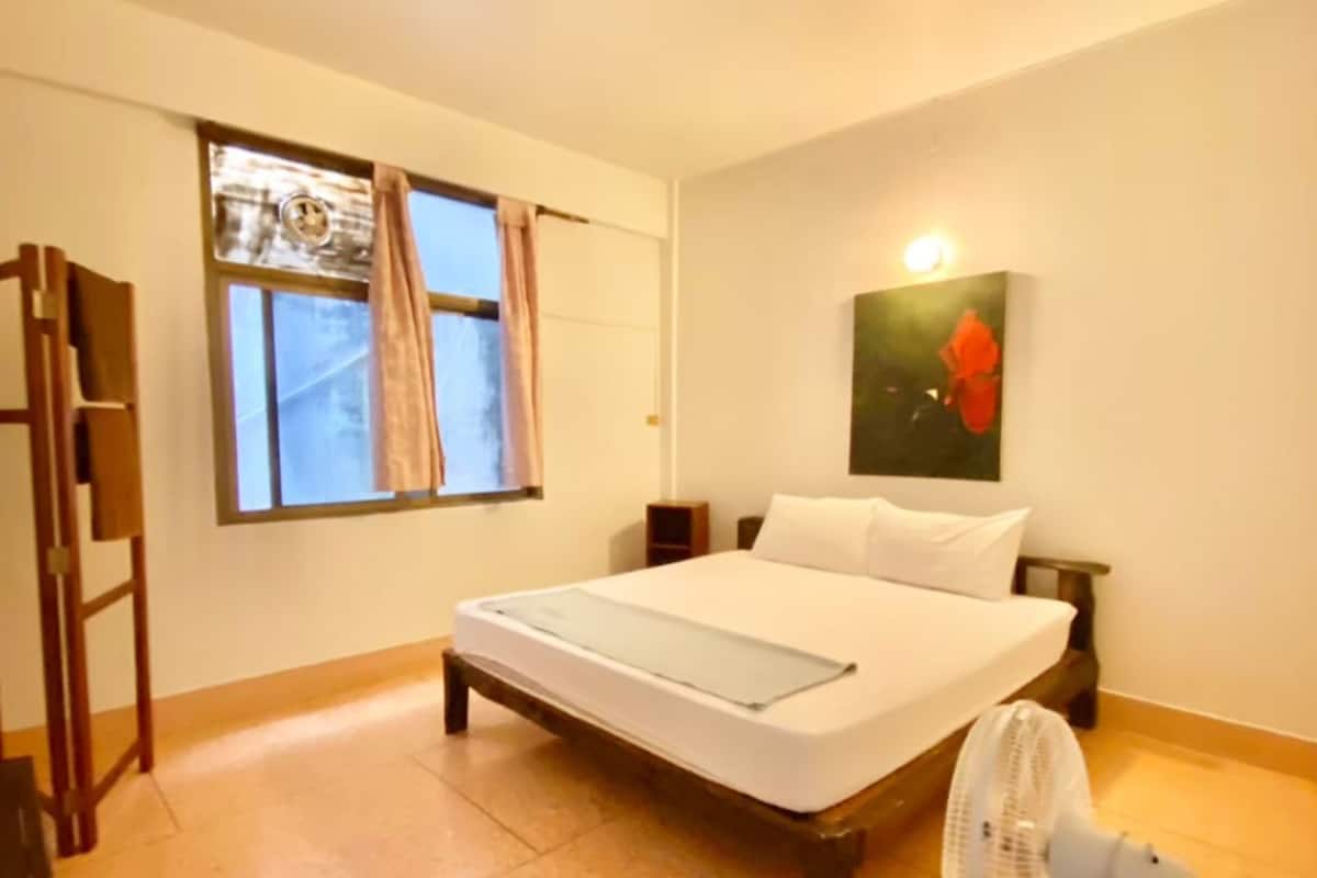 Une chambre à coucher avec un lit, un ventilateur sur pied, une fenêtre avec des rideaux et un tableau accroché au mur.