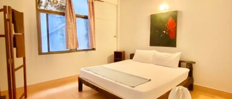 Un dormitorio con cama, ventilador, ventana con cortinas y un cuadro en la pared.