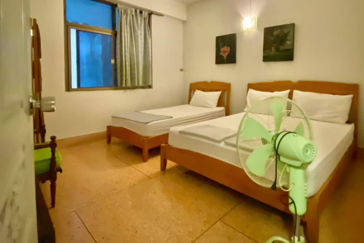 Une chambre avec deux lits simples, un ventilateur sur pied, une fenêtre avec des rideaux et un luminaire fixé au mur.