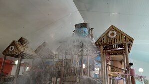 Ein Wasserspielbereich mit verschiedenen Elementen, darunter ein hölzernes Spielhäuschen und eine Rutsche, unter einer weißen Decke.