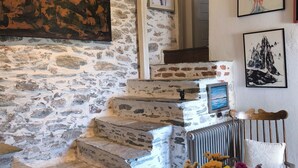 A rustic interior with stone walls, a staircase, a dining table with chairs, and various framed artworks.