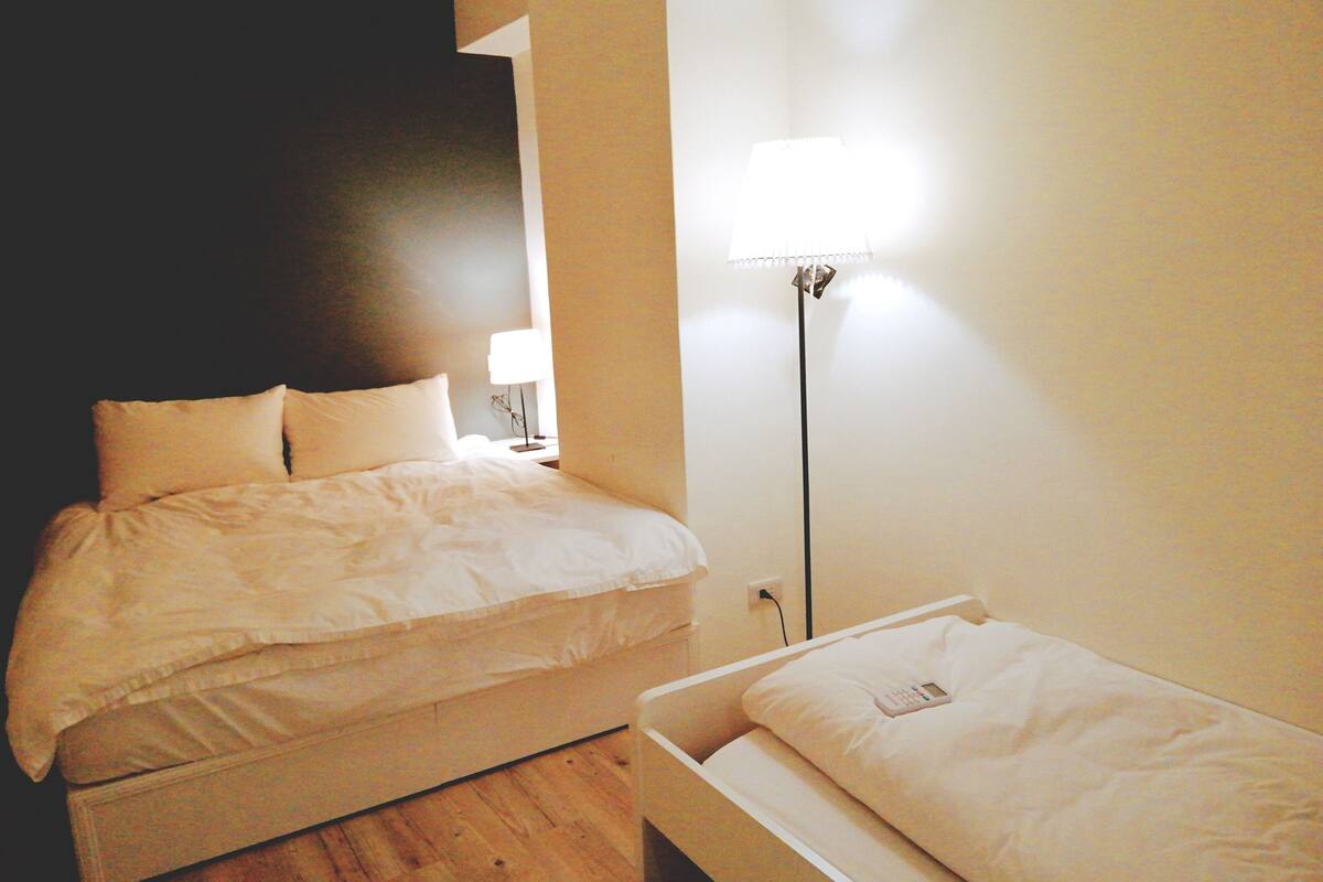 A hotel room with a bed, bedside table, lamp, and a small bedside table with a phone.
