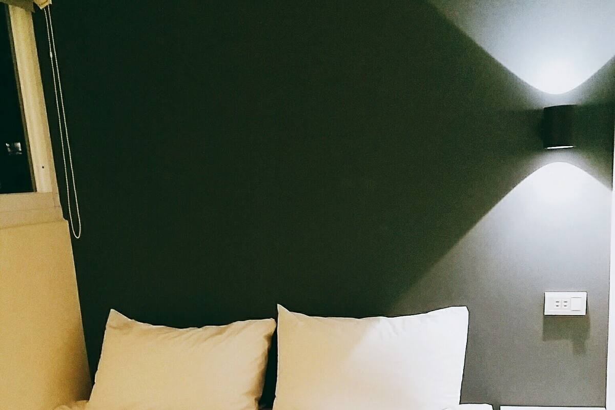 A neatly made bed with white linens in a room with dark walls and a single bedside table.