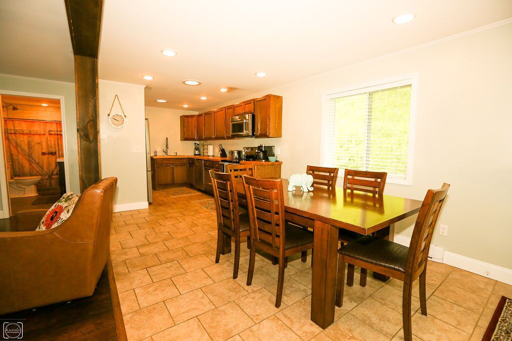 A dining area with a wooden table and chairs, a kitchen in the background, and a window with blinds.