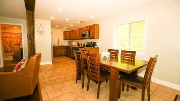 A dining area with a wooden table and chairs, a kitchen in the background, and a window with blinds.