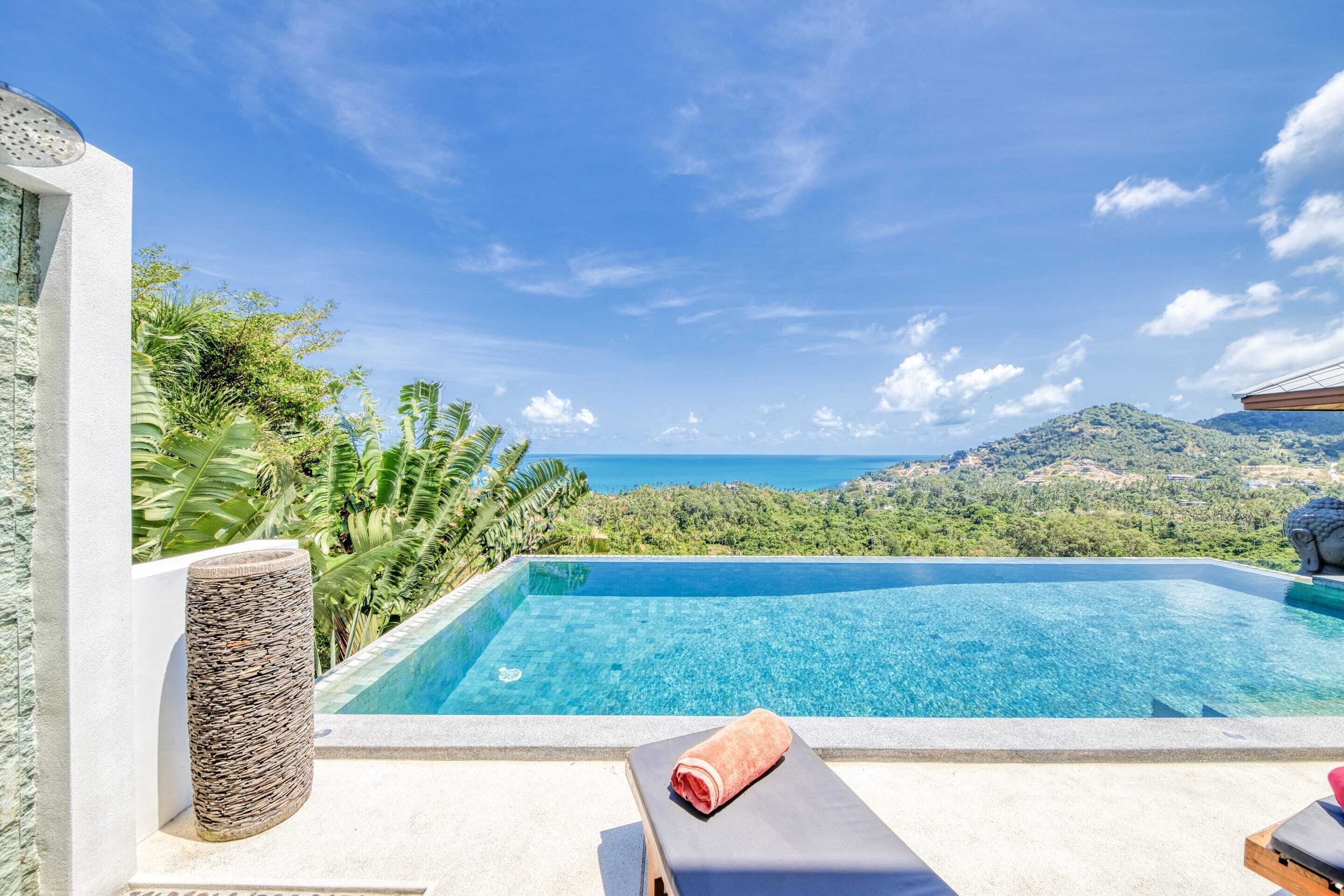 A clear blue swimming pool with a view of the sea and hills, a lounge chair with a towel, and a stone sculpture.