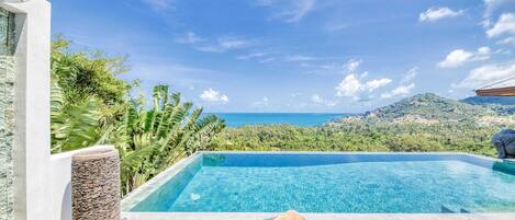 A clear blue swimming pool with a view of the sea and hills, a lounge chair with a towel, and a stone sculpture.