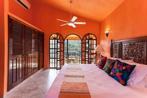 A bedroom with a bed, a ceiling fan, a view through a window, and a door with shutters.