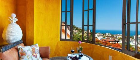A room with a view of a coastal city, a wicker table with a vase and wine glasses, and a sofa with patterned cushions.