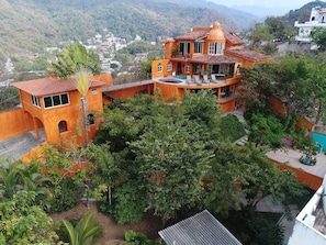 A multi-story building with a balcony, surrounded by lush greenery and a mountainous backdrop.