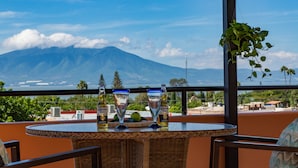 Una terraza con vista a la montaña, una mesa con dos vasos y una botella, y mobiliario de mimbre.