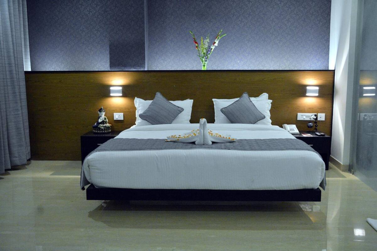 A modern hotel room with a large bed, two bedside tables, a nightstand, a vase with flowers, and a wall-mounted light fixture.