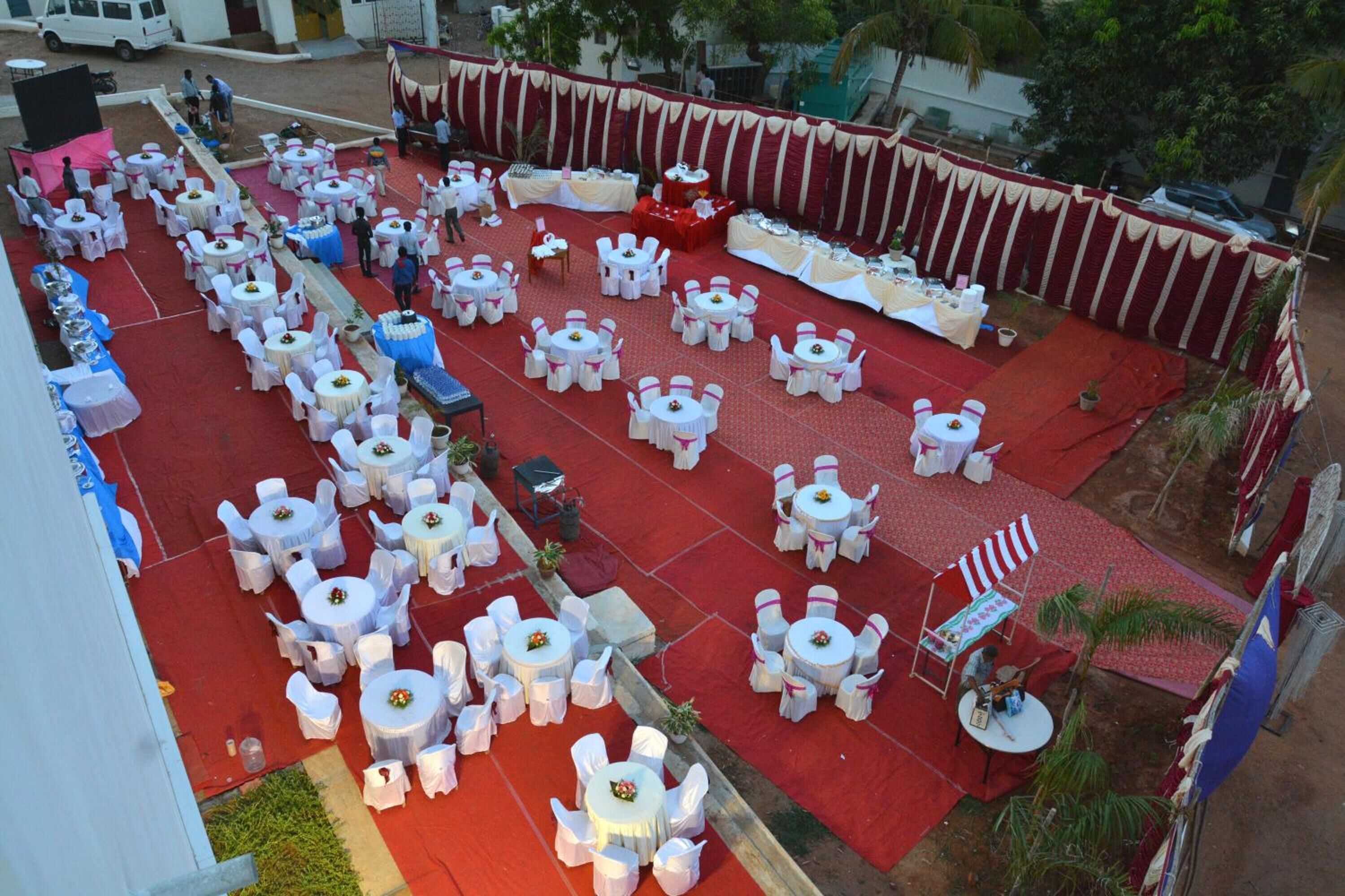 Un espacio para eventos al aire libre con varias mesas redondas y sillas, un escenario con alfombra roja, y una furgoneta blanca al fondo.
