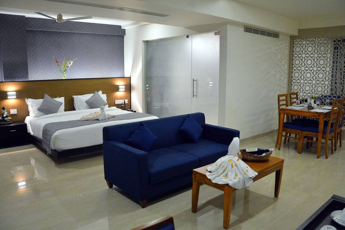 A hotel room with a bed, a blue sofa, a dining table with chairs, and a shower area.