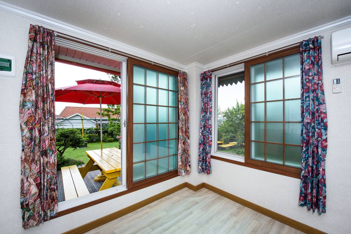 A room with two large windows, each with a wooden frame and frosted glass panes. There are floral curtains on both windows, and an air conditioning unit is mounted on the wall.