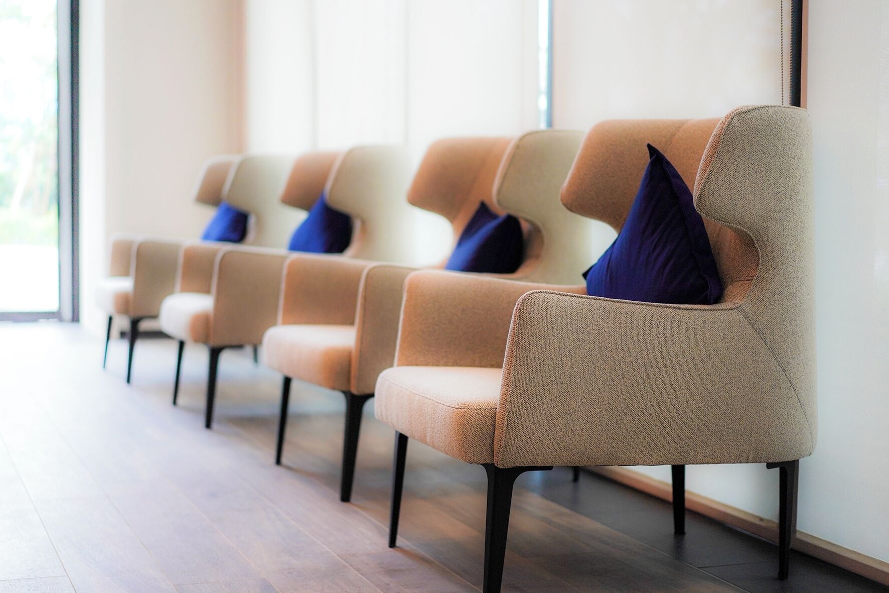 A row of beige chairs with dark legs and blue cushions in a waiting area with wooden flooring and large windows.