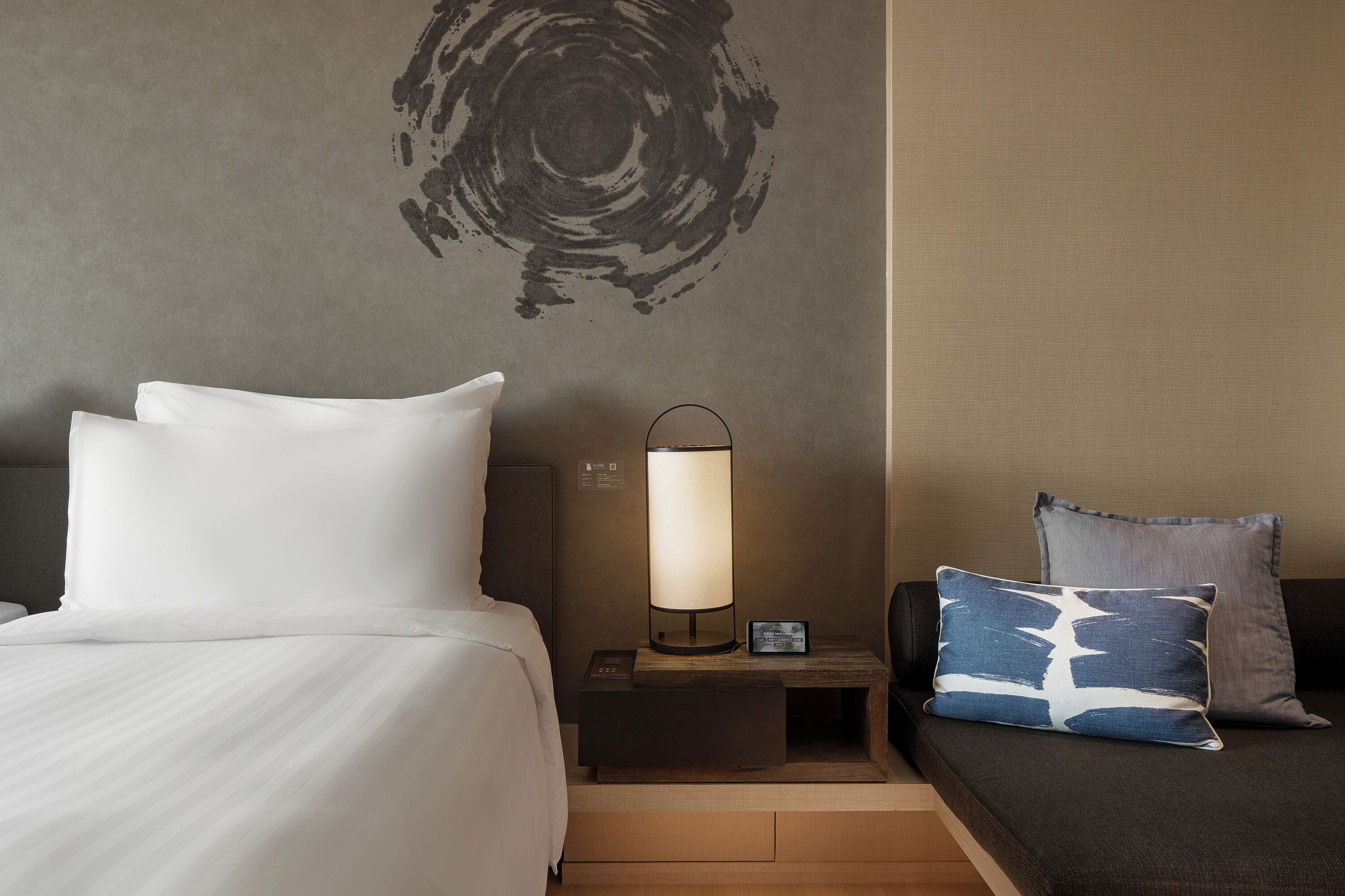 A hotel room with a bed, a bedside table with a lamp, a nightstand, and a sofa with pillows.