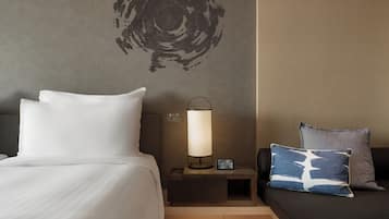 A hotel room with a bed, a bedside table with a lamp, a nightstand, and a sofa with pillows.