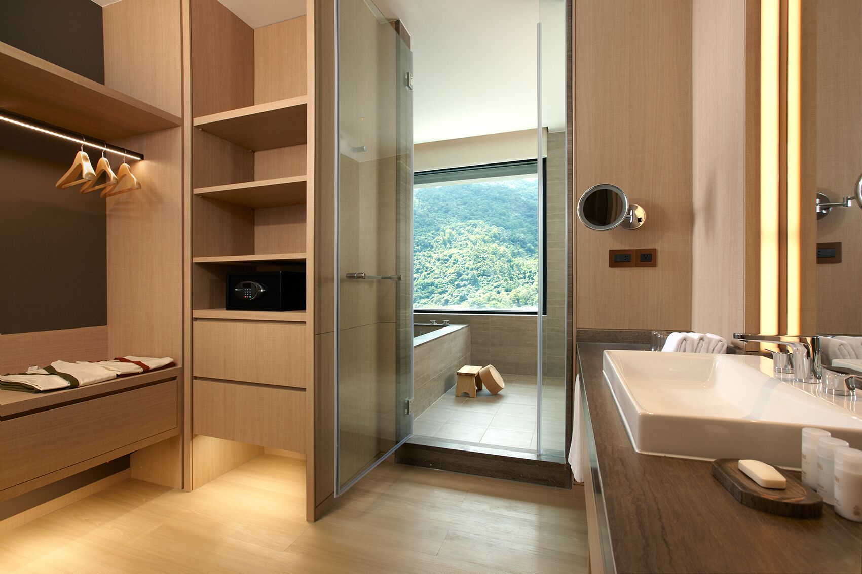 A modern bathroom with a large mirror, a sink, and a shower. There is a view of a mountainous landscape through a glass door.