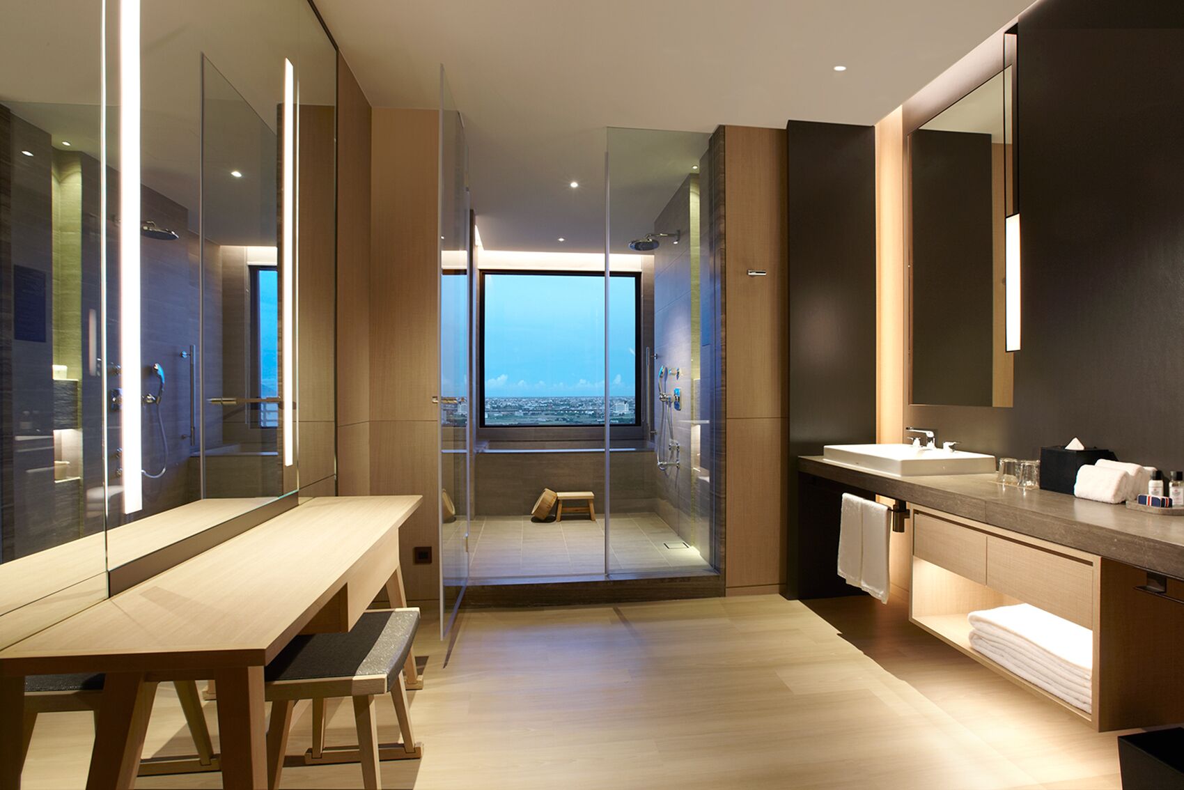 Un baño moderno con ducha envuelta en vidrio, un banco de madera, un espejo grande y vistas a la ciudad.