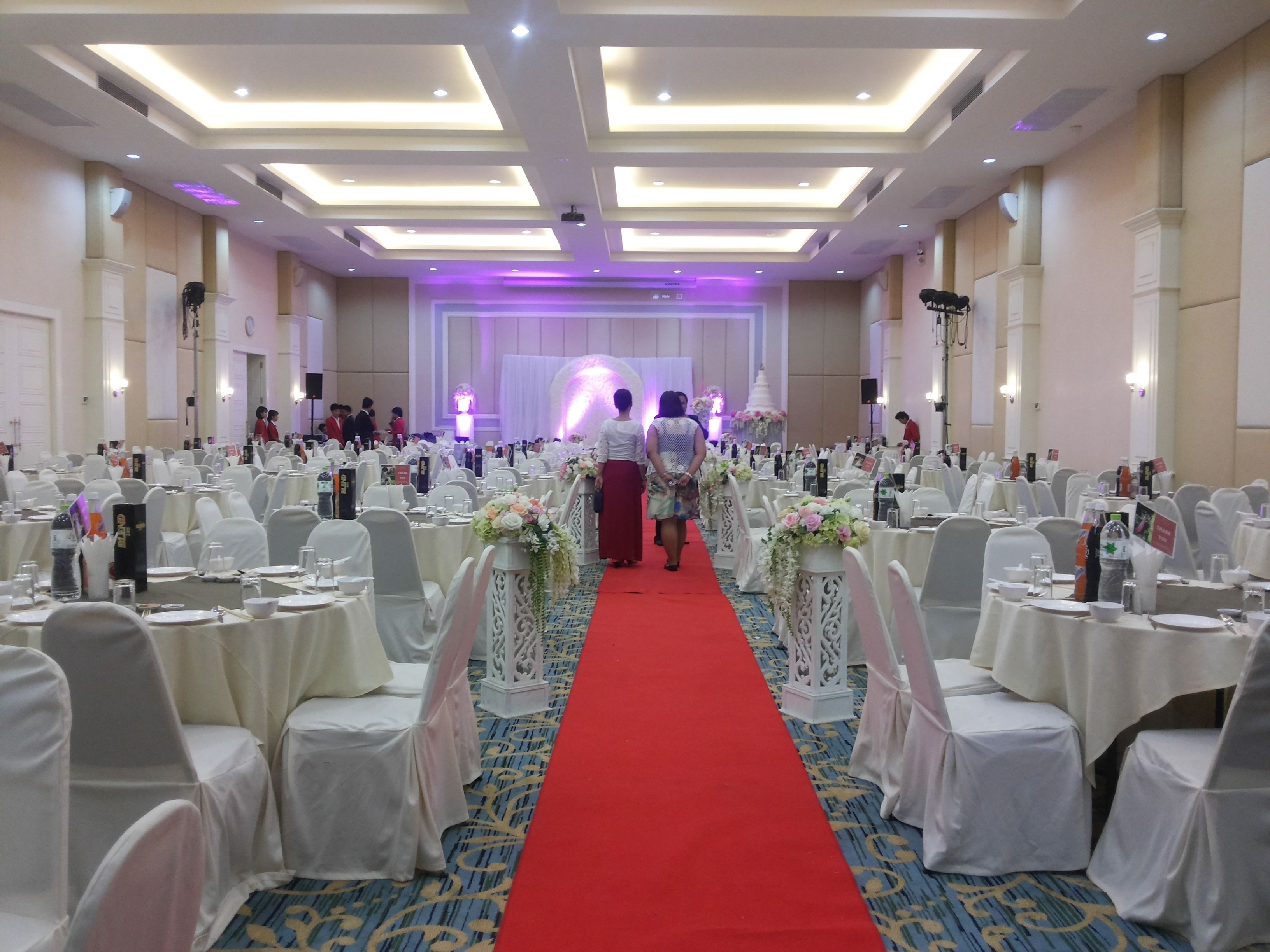 Un salón de banquetes con filas de mesas, sillas blancas y un pasillo con alfombra roja.