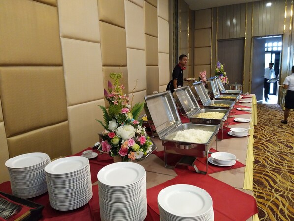 A buffet setup with multiple food stations, a floral centerpiece, and stacked plates and cutlery. - Srilamduan Hotel (Sisaket)