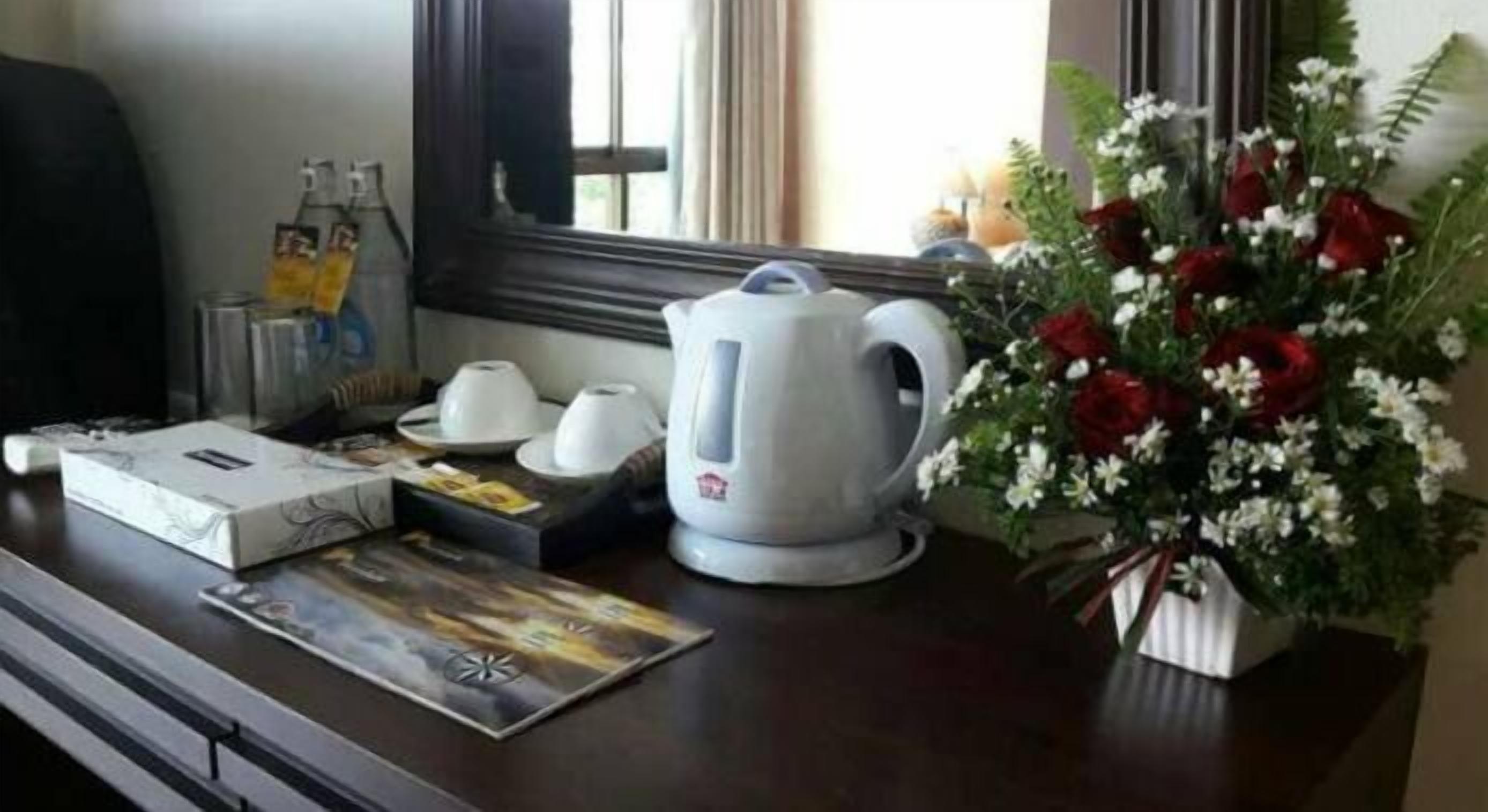 A hotel room with a wooden desk, a white kettle, a bouquet of red and white flowers, and a window with curtains.