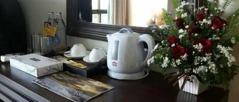 A hotel room with a wooden desk, a white kettle, a bouquet of red and white flowers, and a window with curtains.